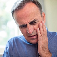 An older man wearing a blue shirt and holding his cheek because he needs to see an emergency dentist in San Antonio