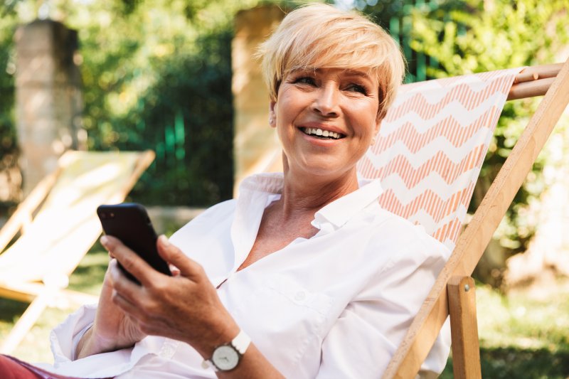 an older woman lounges in a chair in her yard while on her phone and smiling after receiving dental implants in San Antonio