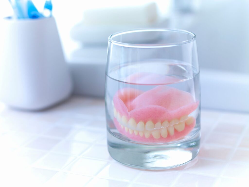 Complete set of dentures in a glass of clear fluid on tiled countertop